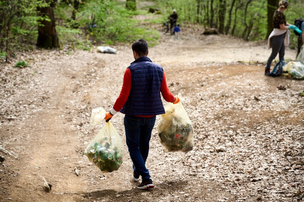 Brian Cristian care participă la o activitate de curățenie, mergând pe un drum de pământ într-o pădure în timp ce poartă două saci mari transparenți plini cu sticle și gunoi colectate. Persoana poartă un tricou roșu cu mâneci lungi, o vestă albastră, blugi și mănuși. În fundal, un alt participant adună gunoiul într-un sac, sugerând un efort de grup pentru curățarea mediului natural.